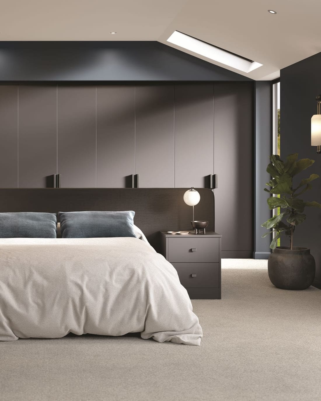 A bedroom with a full wall of floor-to-ceiling fitted wardrobes in dark matt grey, square black handles set in a row, a low panelled headboard running the width of the bed, and matching two-drawer bedside units either side.