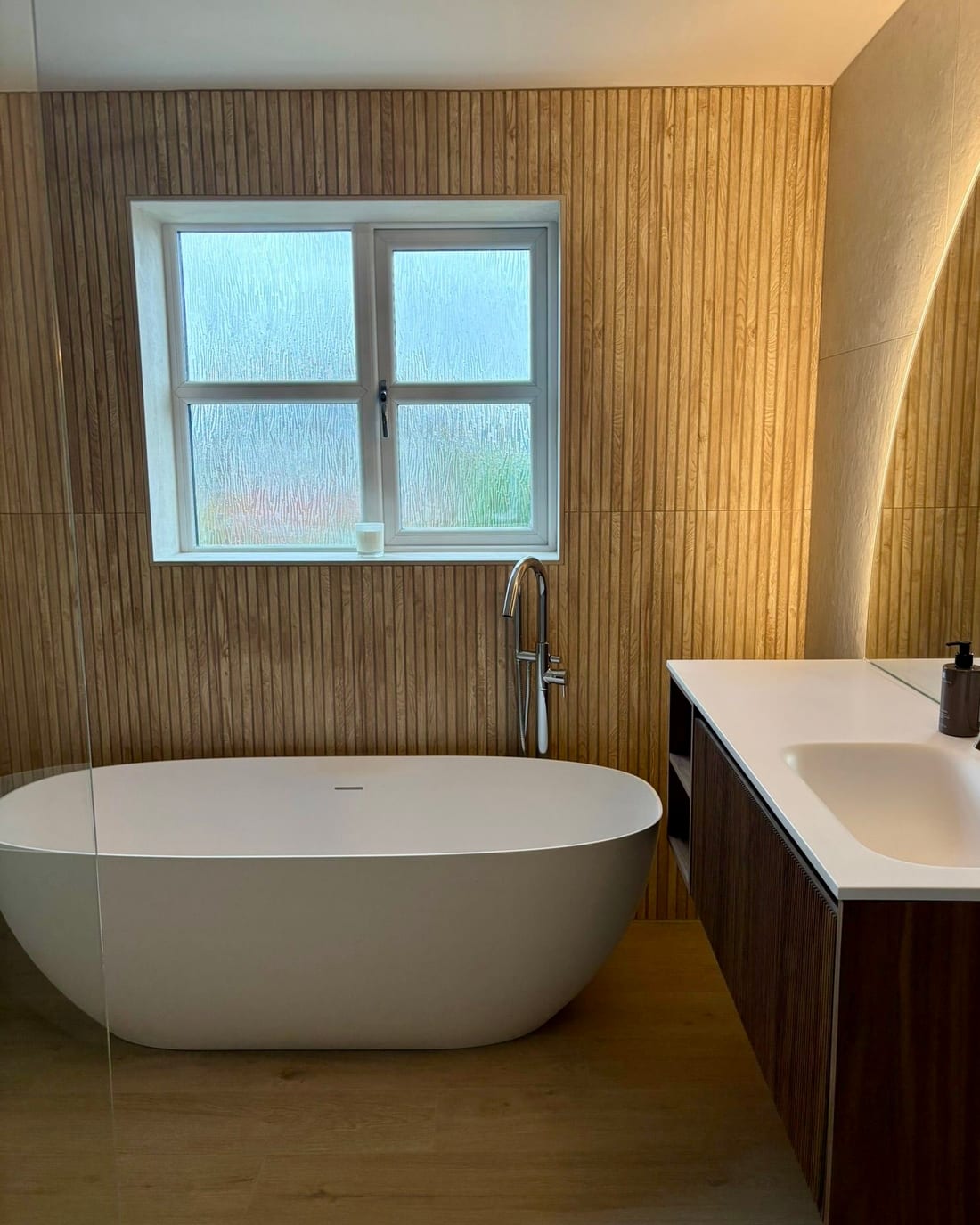 A freestanding white bath beneath a window, with a chrome floor-standing bath filler, fluted oak-effect tiled walls and a walnut wall-hung vanity with a white worktop.
