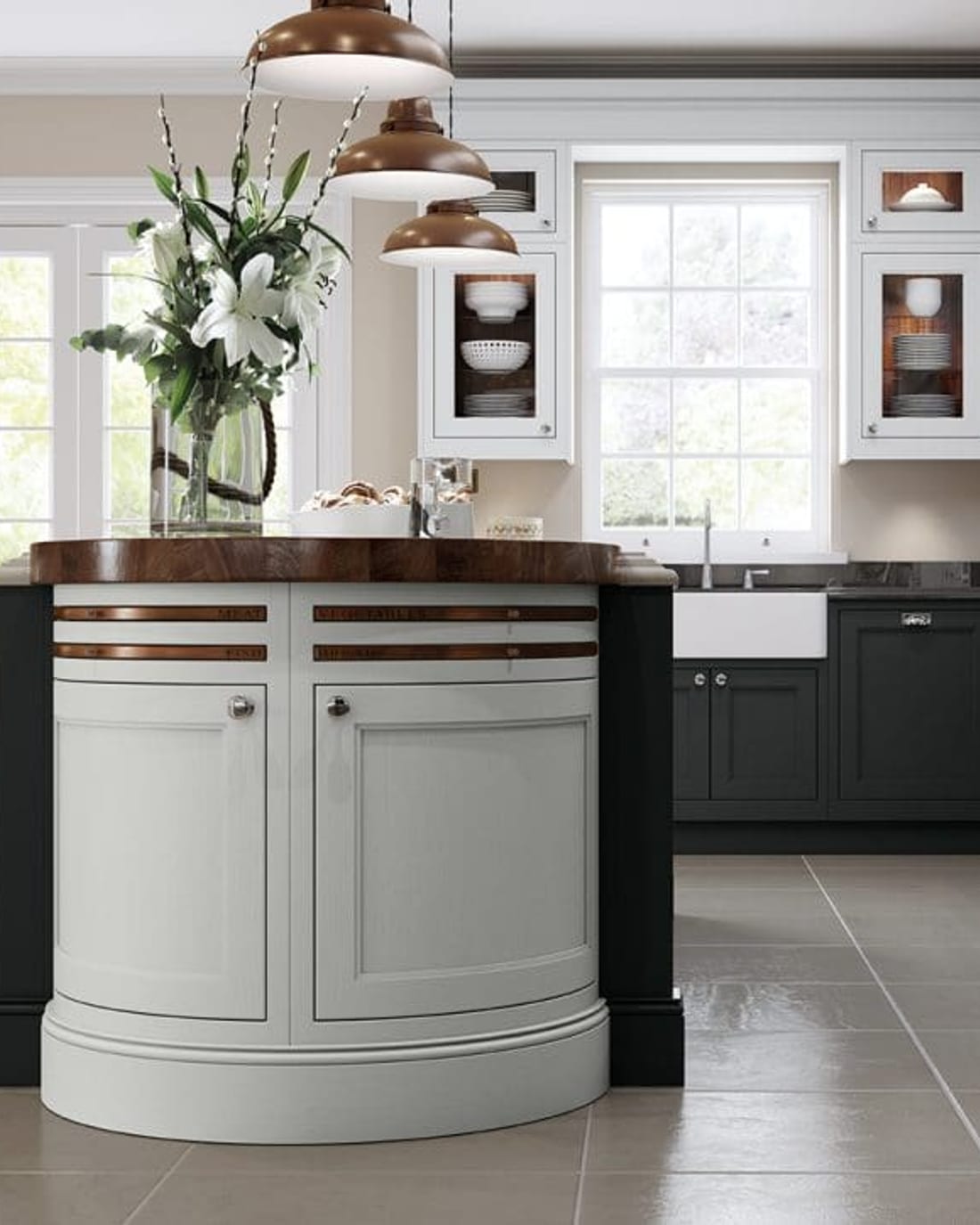 A painted in-frame kitchen with a run of dark grey cabinetry housing a stainless range cooker, and a curved sage-green island beneath copper pendant lights.