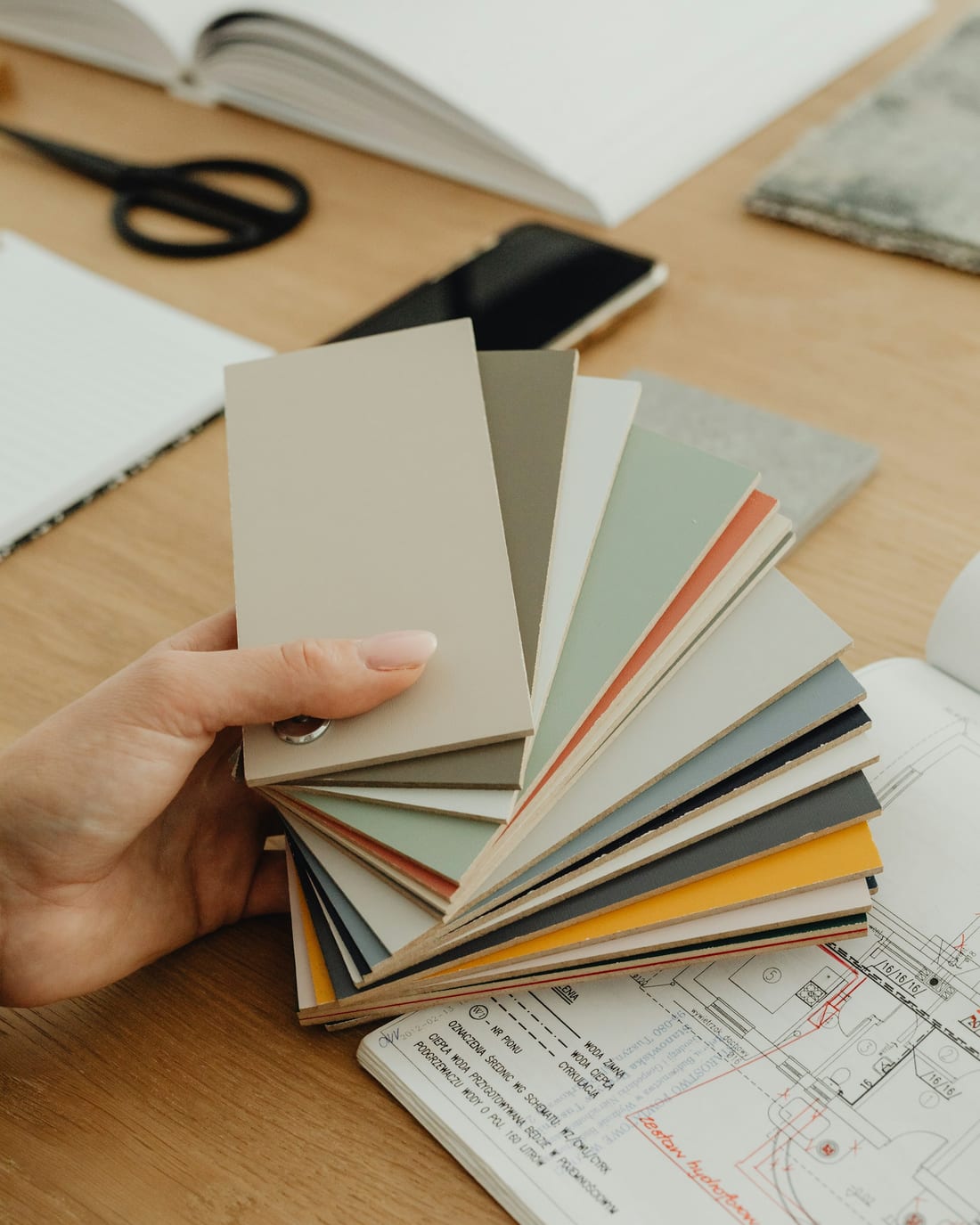 A hand holding a fanned-out set of cabinetry and paint samples in greys, greens and terracotta over a wooden desk, with a technical drawing and a notebook alongside.