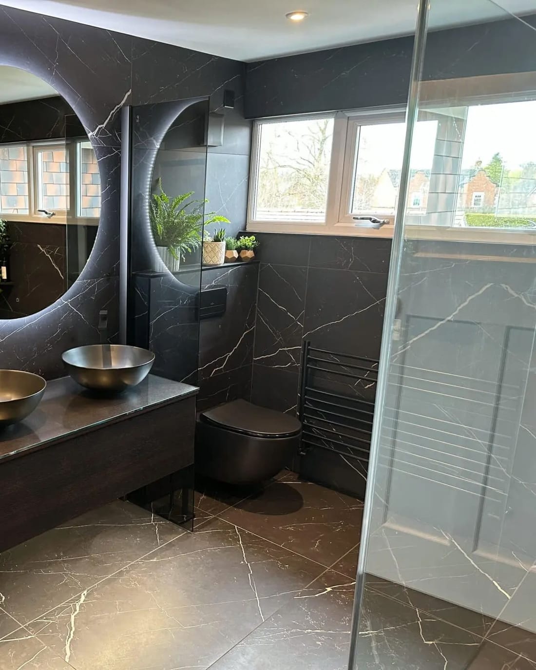 A dark marble-tiled bathroom with a glass shower screen at the right, a wall-hung WC and black heated towel rail against the tiled wall, and twin metal basins on a floating vanity beneath a backlit round mirror.